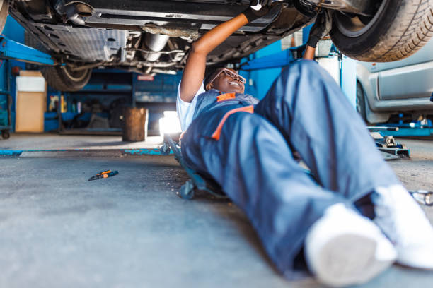 A young African American female mechanic is lying under a car and cheerfully repairing a broken part using a wrench.