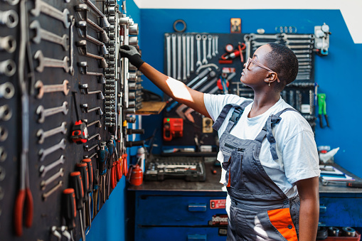 A young African American female mechanic is standing in front of a tool rack and focusing on picking the correct tool.