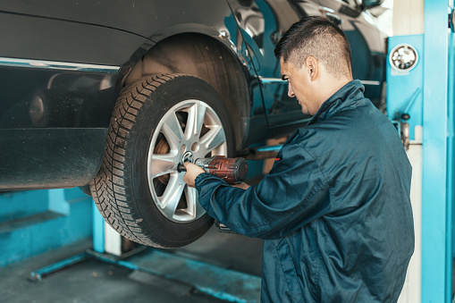 A young Caucasian male mechanic is standing with his back turned and using a drill to tighten bolts on a car tire.