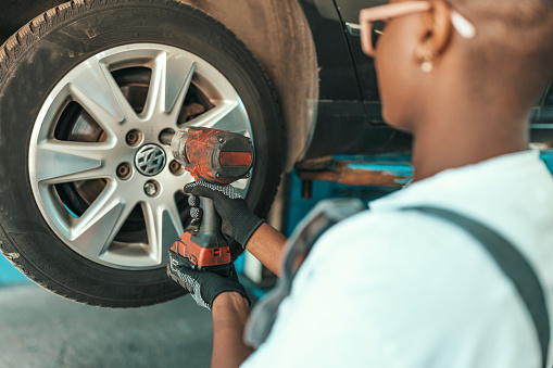 A young African American female mechanic is standing with her back turned and using a drill to tighten bolts on a car tire.