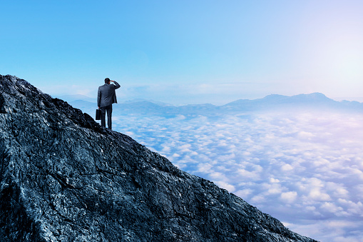 A businessman stands on a mountain top holding his briefcase as he looks out into the distance and the cloud cover below.