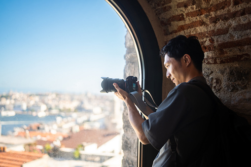 A young male photographer & videographer tourist is taking photos and videos of the city from a window of a historical building during his travel.