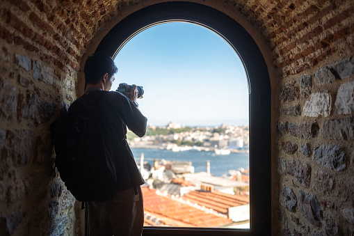 A young male photographer & videographer tourist is taking photos and videos of the city from a window of a historical building during his travel.