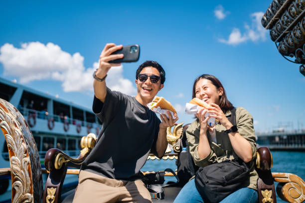 旅行中に屋台の食べ物を体験して食べながらスマートフォンで自撮りをしている観光客のカップル - urban scene women adventure city ストックフォトと画像