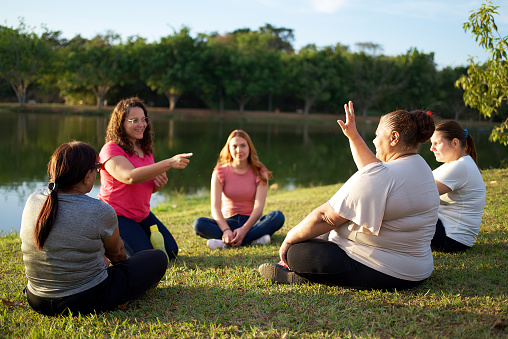 Portrait of a therapy session with some women at a public park, being assisted by a female therapist with conversation techniques. They are all interacting together, talking and having a good moment together, being tenderly cared by their therapist. They are all surrounded by beautiful trees and a lake, with a beautiful lighting from the sun setting.