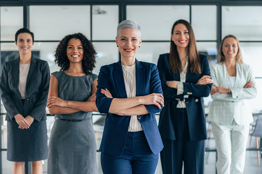 Portrait of beautiful smiling businesswoman with her colleagues. Multiracial  group of businesswomen standing in the office. Successful team leader and her female team in background. Shot of five confident young businesswomen standing in modern office and looking at camera. Group of businesswomen standing together in office.