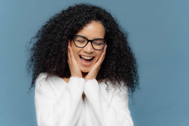 istock Overemotive female touches cheeks laughs from joy, keeps eyes closed, cannot stop giggling, has Afro hairstyle, dressed in white jumper, models against blue background. Ethnicity and emotions concept 1410714922
