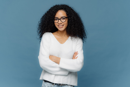 Portrait of delighted pleasant looking curly female keeps arms folded on chest, has confident facial expression, charmin smile on face, wears white sweater and jeans, stands over blue background.