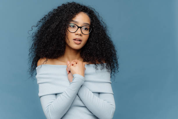 istock Worried nervous female keeps hands pressed together over chest, has thoughtful expression, wears glasses and long sleeved sweater, focused into distance, isolated on blue background with empty space 1410714934