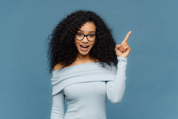 sorprendido mujer feliz con peinado afro, apunta el dedo en el espacio en blanco arriba, ha abierto la boca, lleva gafas y jersey con los hombros desnudos, aislado en la pared azul. concepto publicitario - frizzy 20s adult african descent fotografías e imágenes de stock