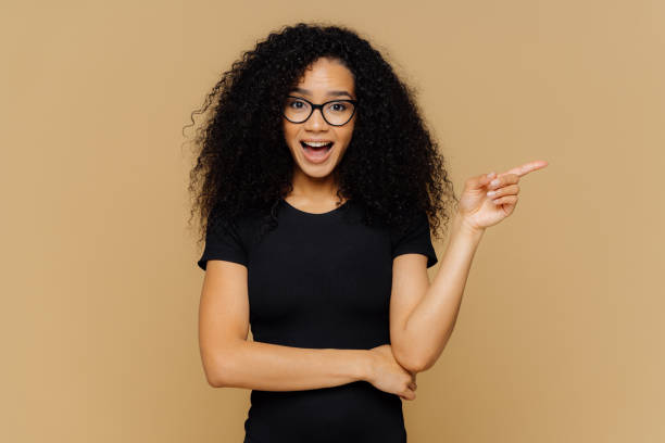 istock Brunette mixed race woman points aside with index finger, has glad curious expression, promots awesome product, dressed in casual clothes, isolated over brown background. People and advertisement 1410714965