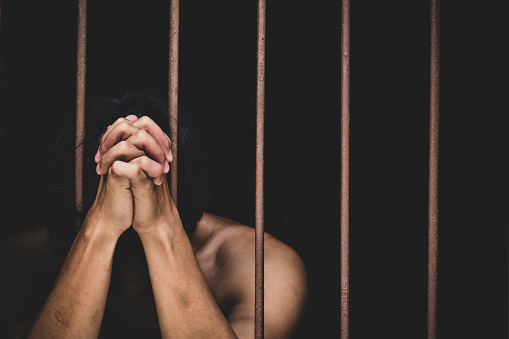 Female prisoner in orange uniform holds hands on metal bars, walks, looks at barred window in jail cell. Woman serves imprisonment term for crime in prison. Depressed inmate in correctional facility.
