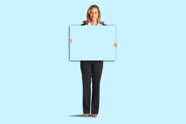 A businesswoman smiles and looks directly at the camera as she holds a blank blue card in front of a blue background of the same color.