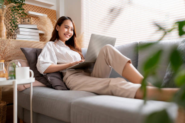 young attractive asian female working at home hand use laptop checking search schedule working process planning report with leisure relax on sofa in living room with morning light peaceful moment stock photo