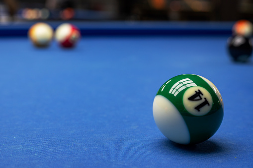 The Fourteen Ball on a pool table with other balls in background. Fourteen Ball is in focus while the others are heavily out of focus. Pool table is lit by an overhead light and is blue.