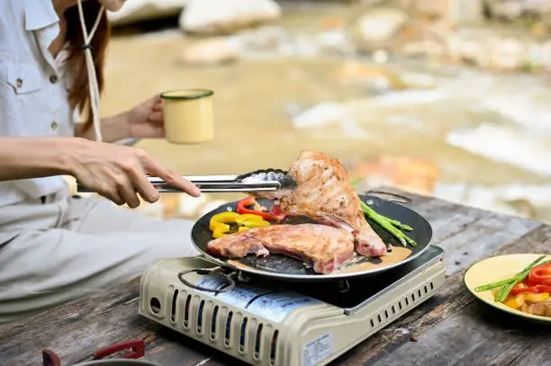 Attractive young Asian female camper grill some pork-chop steaks on a camping picnic stove near the river. Camping concept. cropped and close-up image