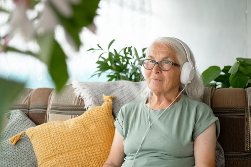 Caucasian elderly gray haired woman with headphones is sitting at home on couch and listening to music, audiobook, lecture. Concept of audio healing and sound therapy, learning