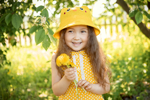 Summer portrait of cute beautiful smiling Caucasian girl in yellow hat and dress in park with bouquet of yellow dandelions in her hands