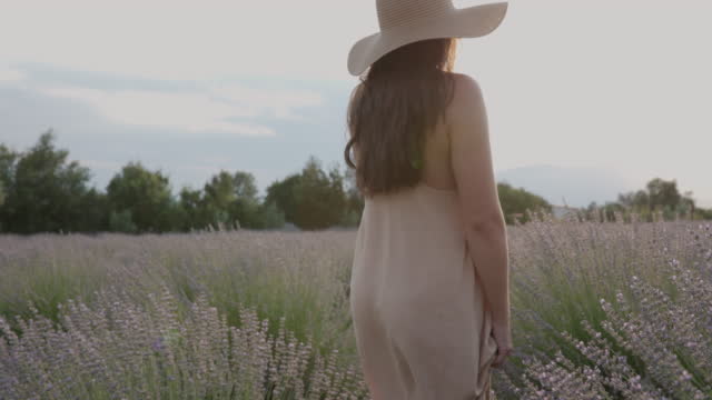 Young woman in summer dress and sun hat walking through lavender field at sunset