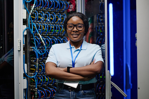 Portrait of African female engineer smiling at camera standing with her arms crossed in data server with cables in rack in background