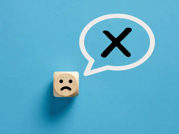 Photo of Unhappy face icon on a wooden cube with cross x or no sign in a speech bubble. No or rejection