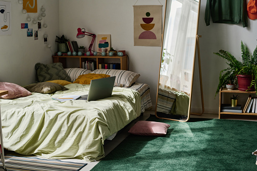 Horizontal image of modern teenage bedroom with bed and laptop on it with big mirror in centre of the room