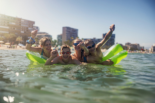 Mother and teenage kids are are having fun on inflatable pool raft is sea. Safe, shallow sandy beach.\nSunny summer day in Alicante, Spain.\nCandid smile and laughter. Shallow DOF. Family is cheering and laughing at the camera.\nShot with Canon R5.