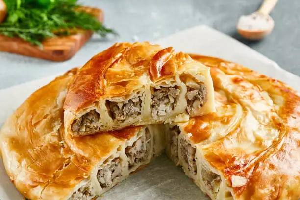 Photo of Classic turkish pie with meat on wooden board. Composition with burek pie on concrete background with textile and spices. Balkan pie with minced meat  in rustic style on gray table.