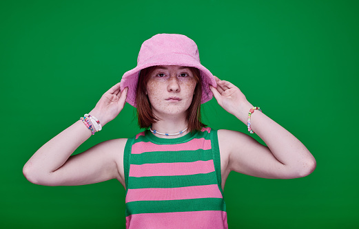 Portrait of teenage girl in pink panama hat looking at camera standing against green background