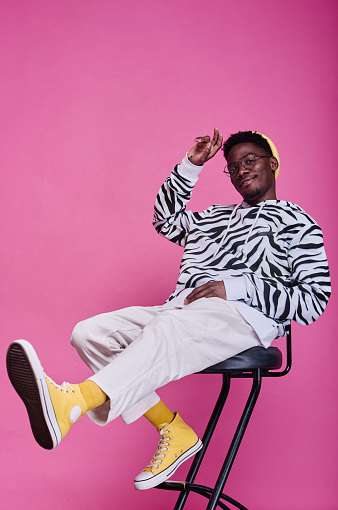 Portrait of African teenage boy in stylish clothes sitting on chair and posing at camera against pink background