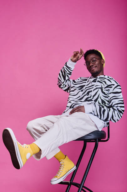 African teenage boy in stylish clothes Portrait of African teenage boy in stylish clothes sitting on chair and posing at camera against pink background cool attitude fashion stock pictures, royalty-free photos & images