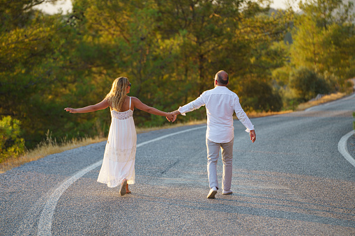 Portrait of cheerful mid adult couple embracing, holding hands walking on the road at sunset