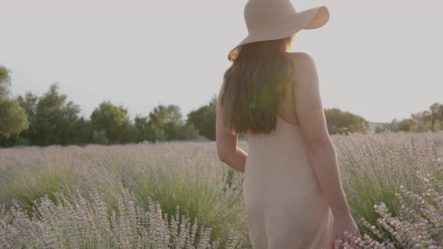 Young woman in summer dress and sun hat walking through lavender field at sunset