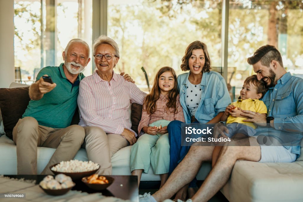 Three generation family sitting on the sofa and watching TV Multi-generation family with a TV remote control in the living room. Front view of a family watching TV on the sofa at home. Grandpa holding a tv remote controlor. Watching TV Stock Photo