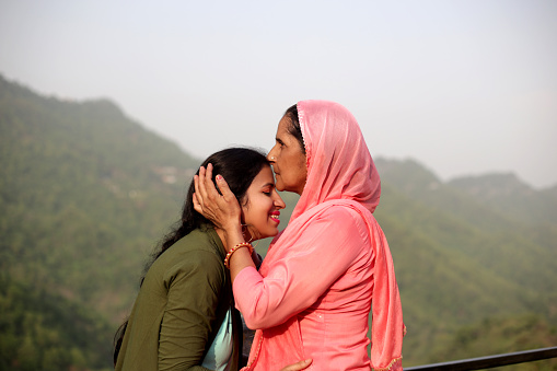 Mother hugging her daughter and kissing her outdoors against mountain background.