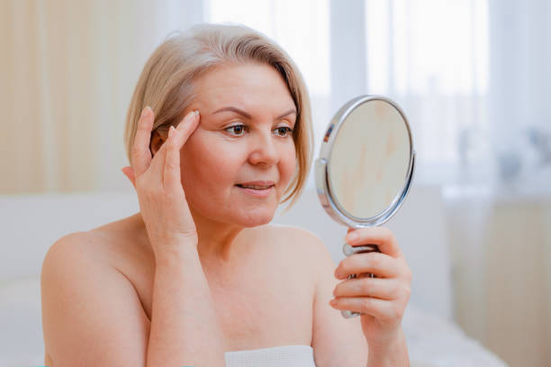 portrait pretty senior woman with hands on her face mirror at home after bathroom skin care after 50-60 years - make up women ceremonial makeup senior women imagens e fotografias de stock