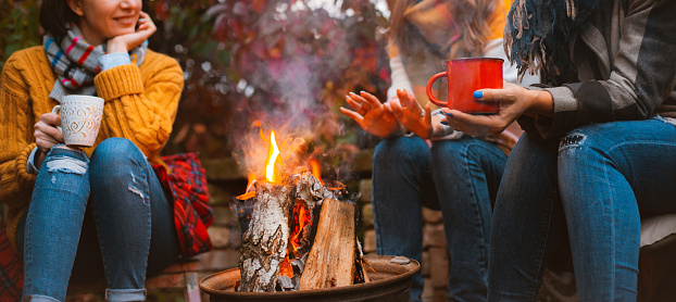 Cropped photo of three females best friends sitting around bonfire in casual clothes warming up and communicating, holding mugs with hot drinks, happy to be together outside in cold autumn evening