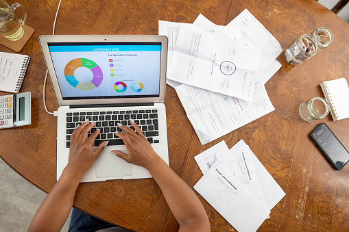 A view looking down from above of an unrecognizable person typing into the laptop and going through personal finances. The table is covered in bills and various documents.