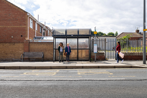 A mid-female adult walks to meet her sister and mother at the bus stop. Her sister and Grandmother are waiting patiently, all wearing casual clothing.