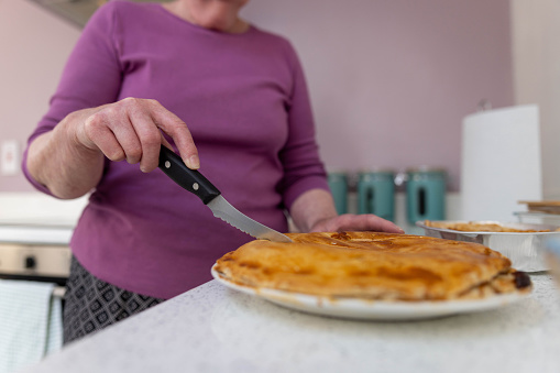 Close up of unrecognisable senior lady cutting into a freshly cooked pie. The lady is wearing a vibrant purple top