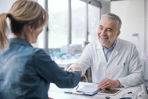 Smiling doctor sitting at desk and giving a handshake to his patient, medicine and healthcare concept