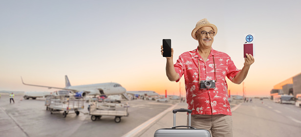 Mature male tourist with a suitcase showing a passport and a smartphone on an airport apron