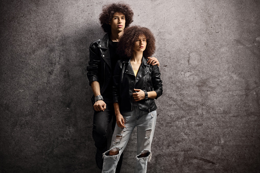 Young man and woman with leather jackets and curly hair leaning on a dark background