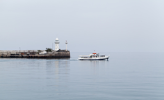 Coastal view of Yalta port. Small passenger ferry ship sails near white lighthouse tower on a cloudy day