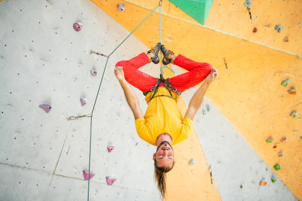A man hangs upside down in a lotus position on a climbing wall. The man is wearing a yellow T-shirt and red pants.