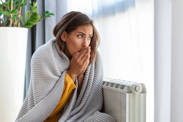 woman freezing at home, sitting by the cold radiator. woman with home heating problem feeling cold - tremendo imagens e fotografias de stock