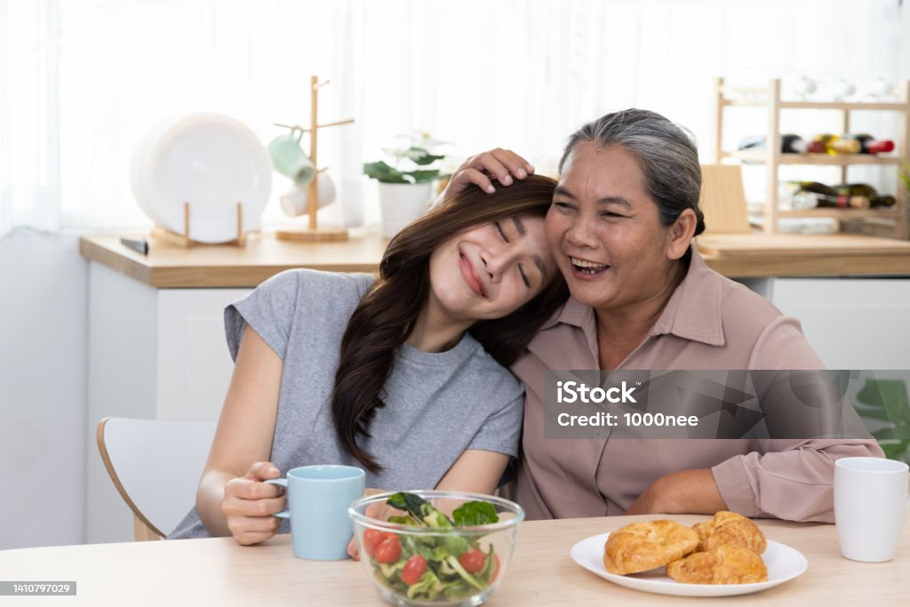 Happy Asian mature mother and daughter laughing and embracing Happy Asian mature mother and daughter laughing and embracing at the breakfast table in the kitchen Mother Stock Photo