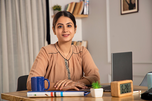 A Happy Smiling attractive confident motivated Asian Modern Indian Woman or female or formal start up corporate office lady employee or businesswoman sitting at workplace and looking at the camera.
