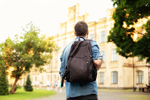 Student university standing with his back to the camera and his backpack on one shoulder and walking in university campus, education concept. Young man walking down street with a backpack. Back view.