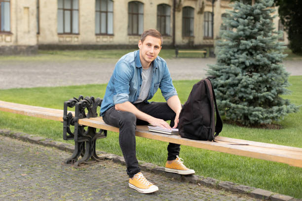 istock Young man reading book while sit on bench at university campus. Happy Man study remotely. Adult student reading in the park. Enjoying student life. smiling man working on outdoor. education concept 1410805822